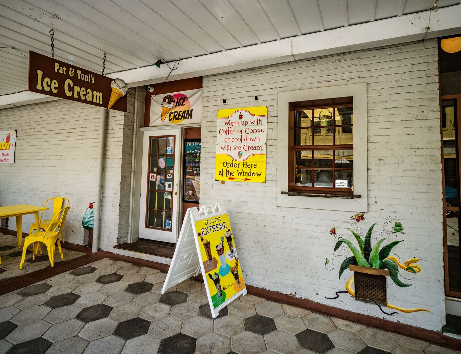 Small ice cream shop storefront with yellow chairs and sandwich board signs on a tiled walkway.