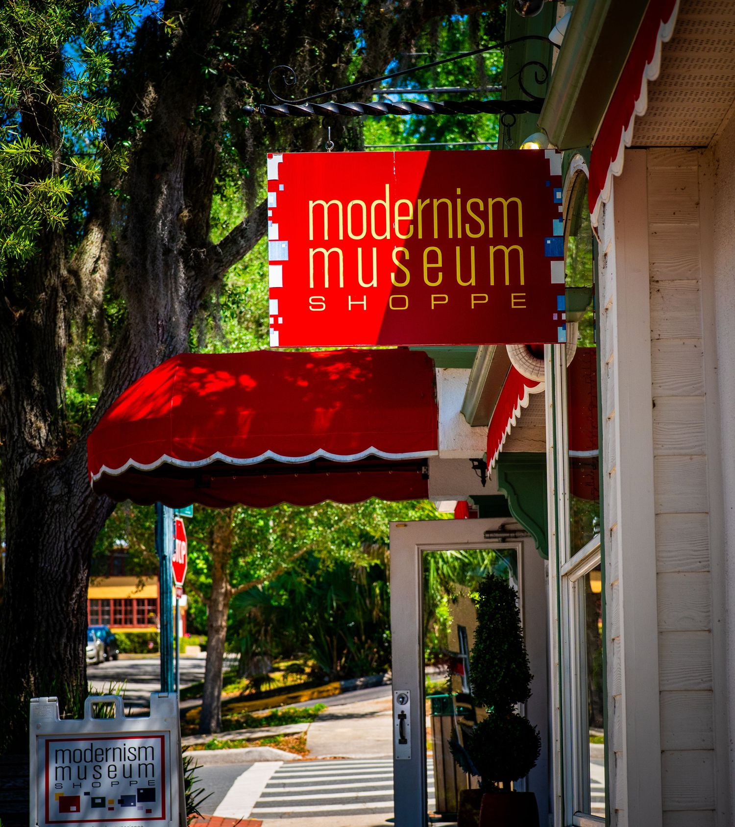 Red modernist shop awnings against a bright blue sky with lush green trees in the background.