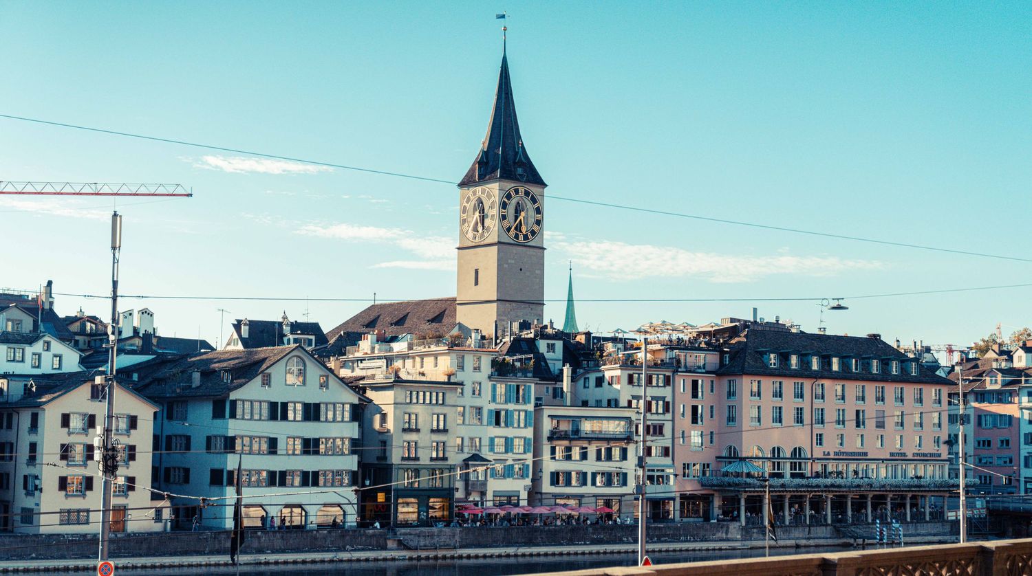 Historic European old town skyline with a tall clock tower rising above colorful buildings along a riverfront under a clear blue sky.