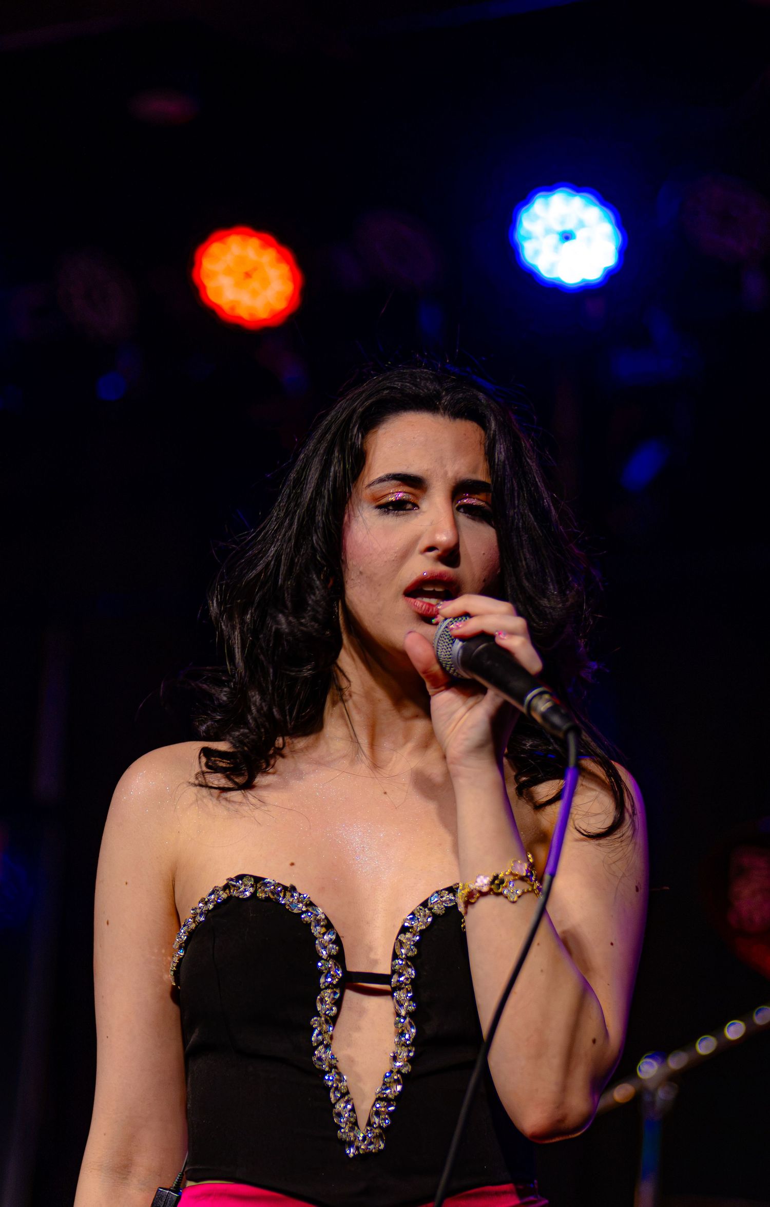 Female singer performing on stage in black jeweled strapless top, holding microphone under colorful stage lights.