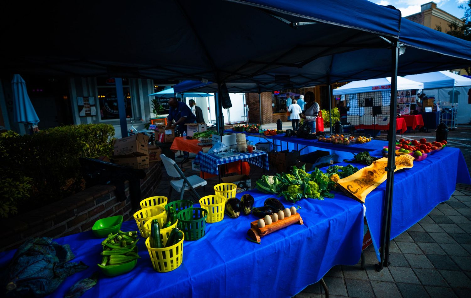 Fresh produce and goods displayed on blue-covered tables at an outdoor farmers market at dusk.