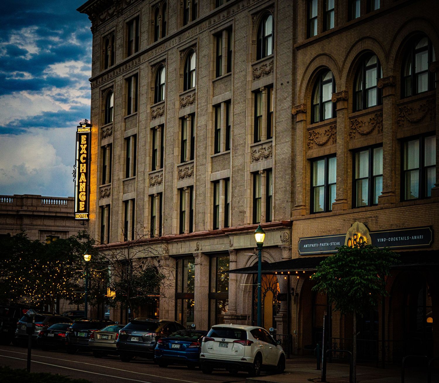 Historic ornate stone hotel building with arched windows, glowing street lamps, and parked cars at dusk in downtown.