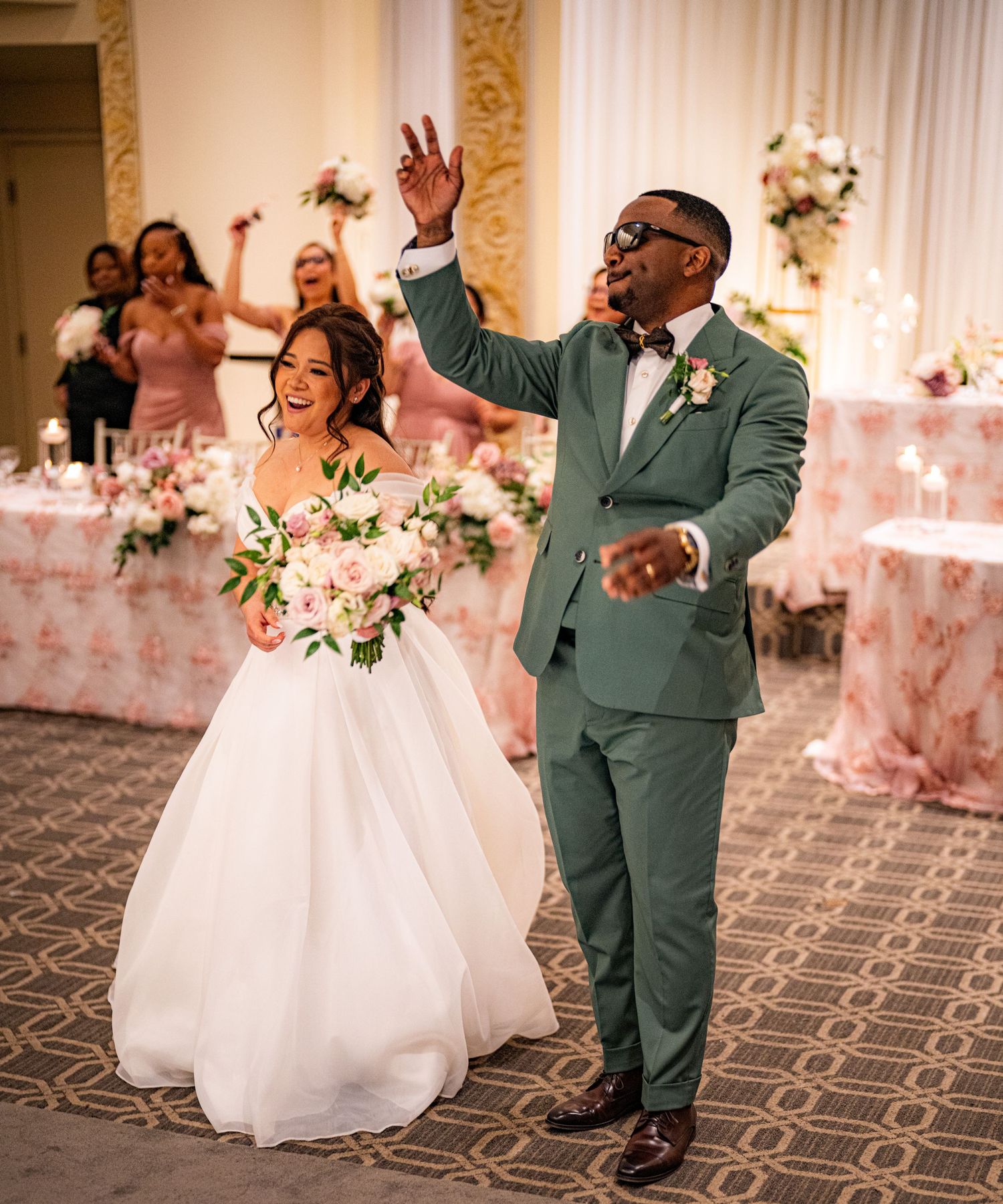 Bride in white ball gown and groom in green suit celebrate their wedding reception entrance with raised hands.