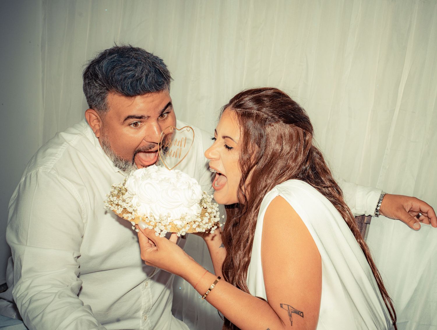A couple shares playful moments while smashing cake in each other's faces during a wedding celebration.