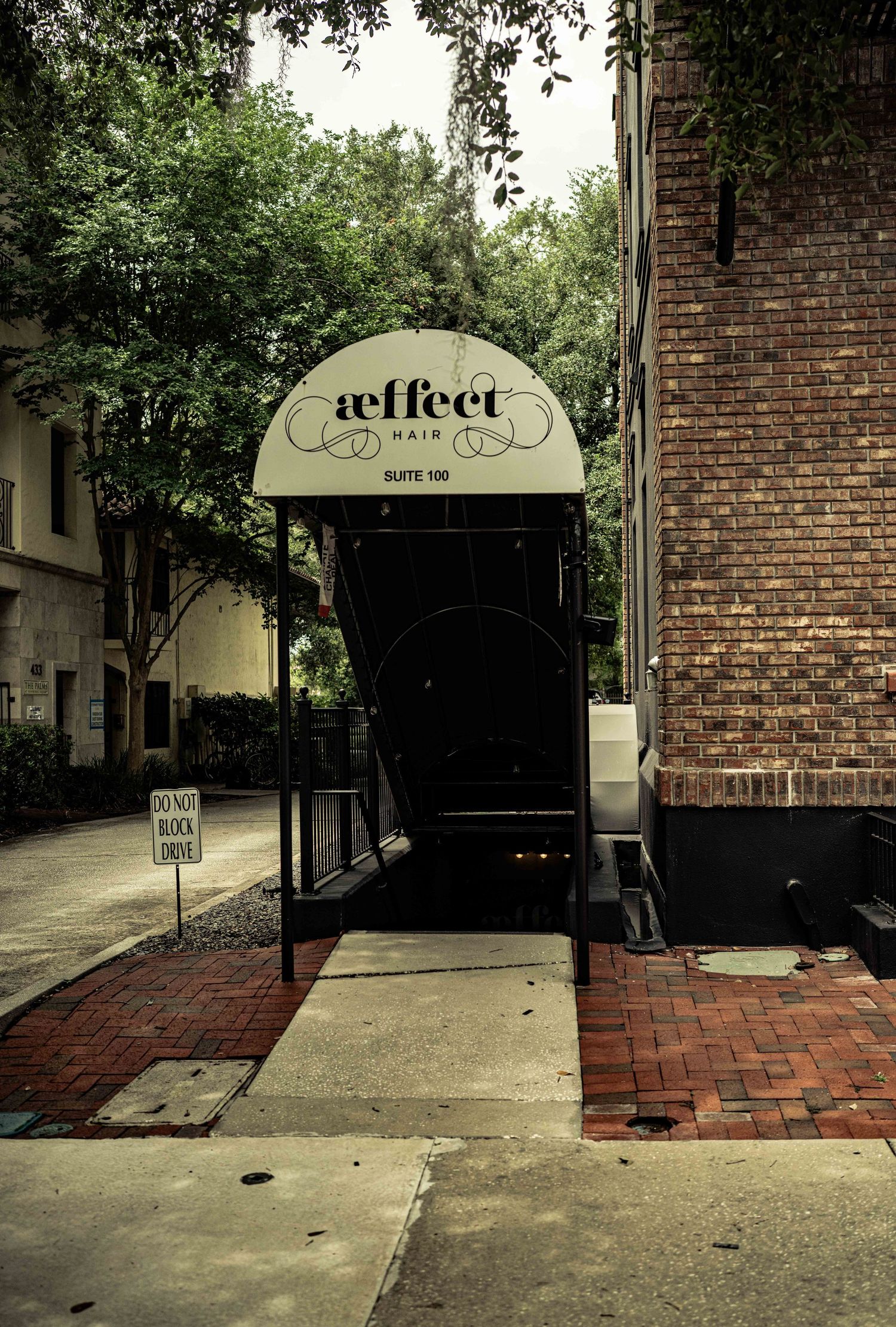 Aeffect Hair salon entrance with elegant awning at Suite 100, framed by brick walls and lush trees.