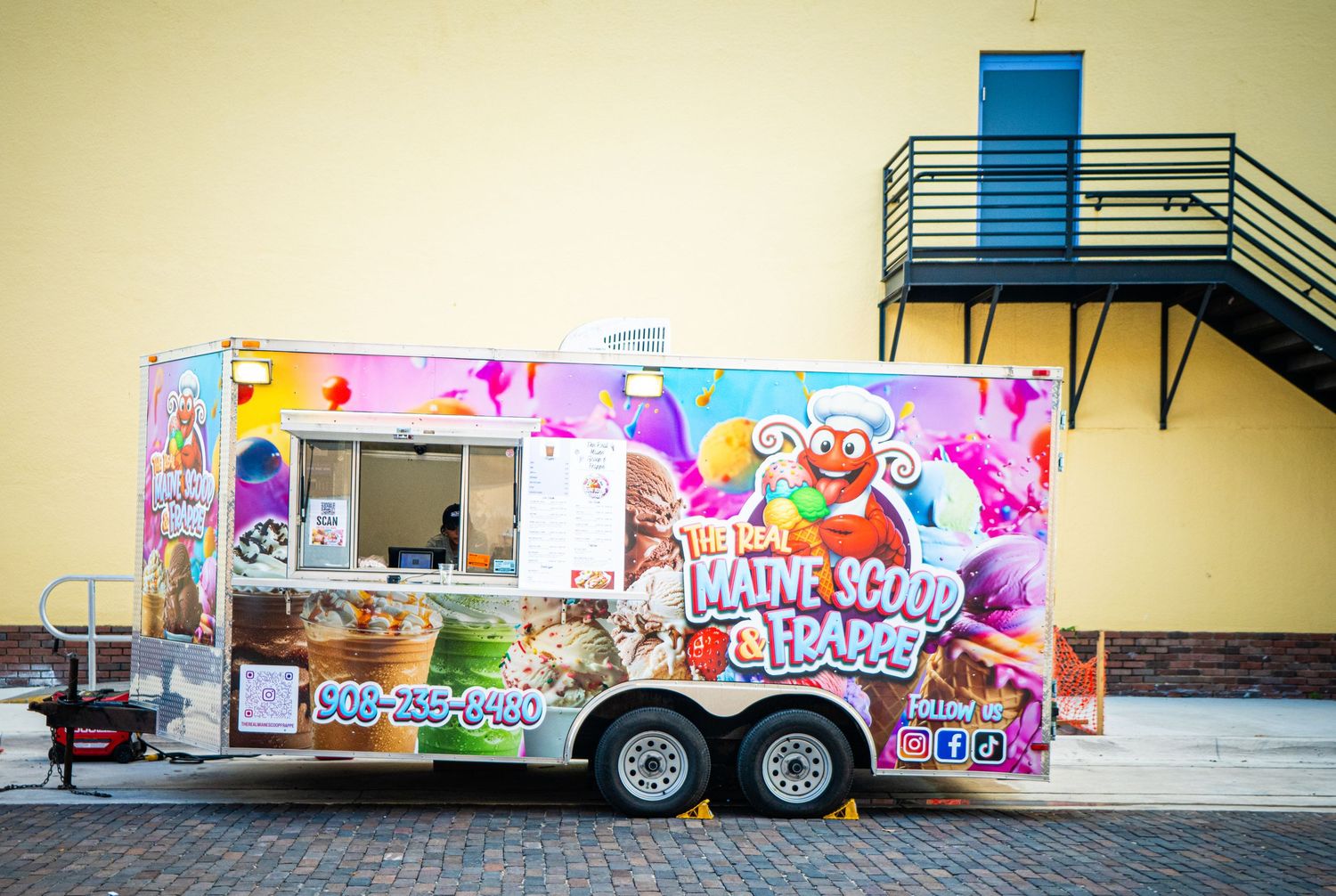 A colorful food truck with cartoon designs and vibrant pink decorations parked against a yellow building wall.