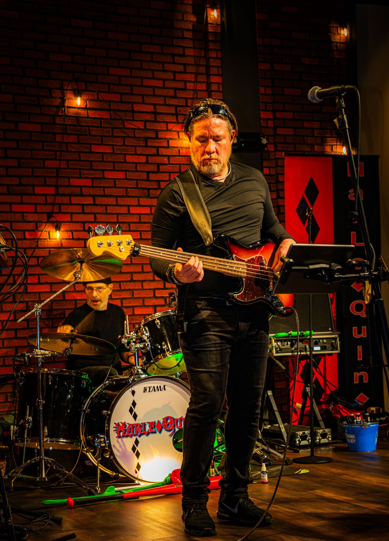 Bass player performs on stage with red brick wall and drum kit in background.