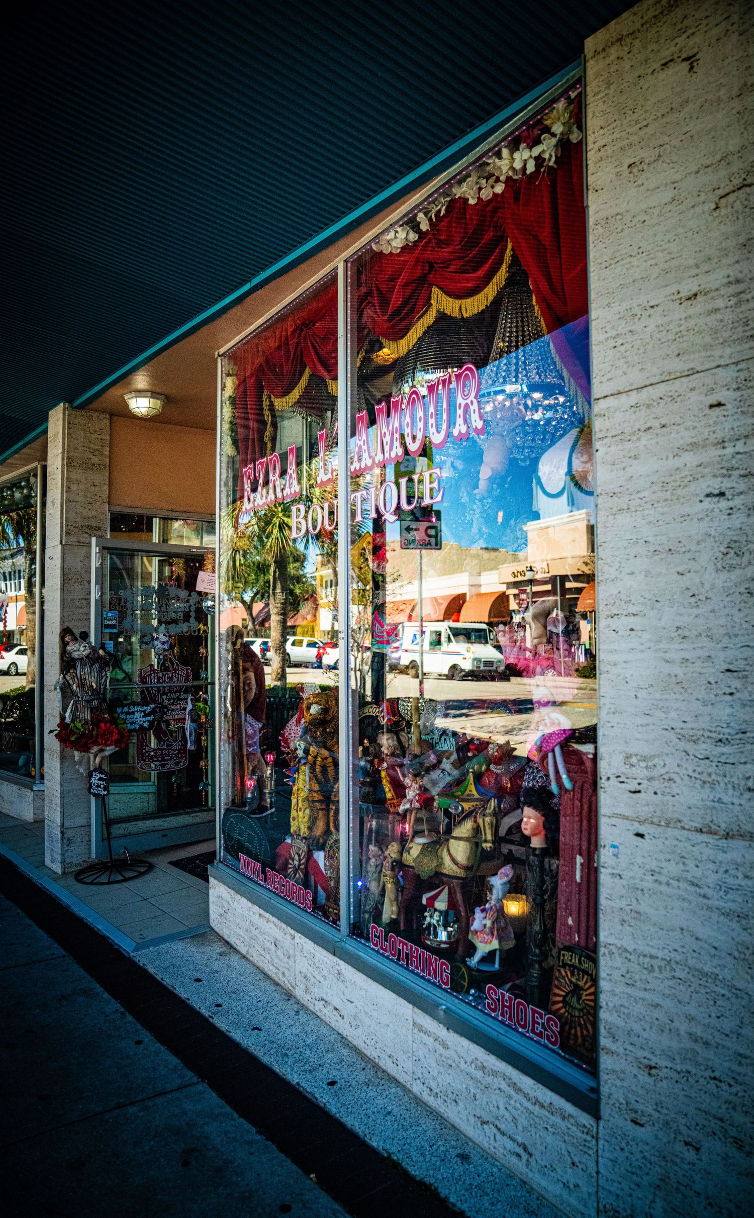 Retail storefront windows displaying colorful clothing and merchandise with a white brick exterior in evening light.