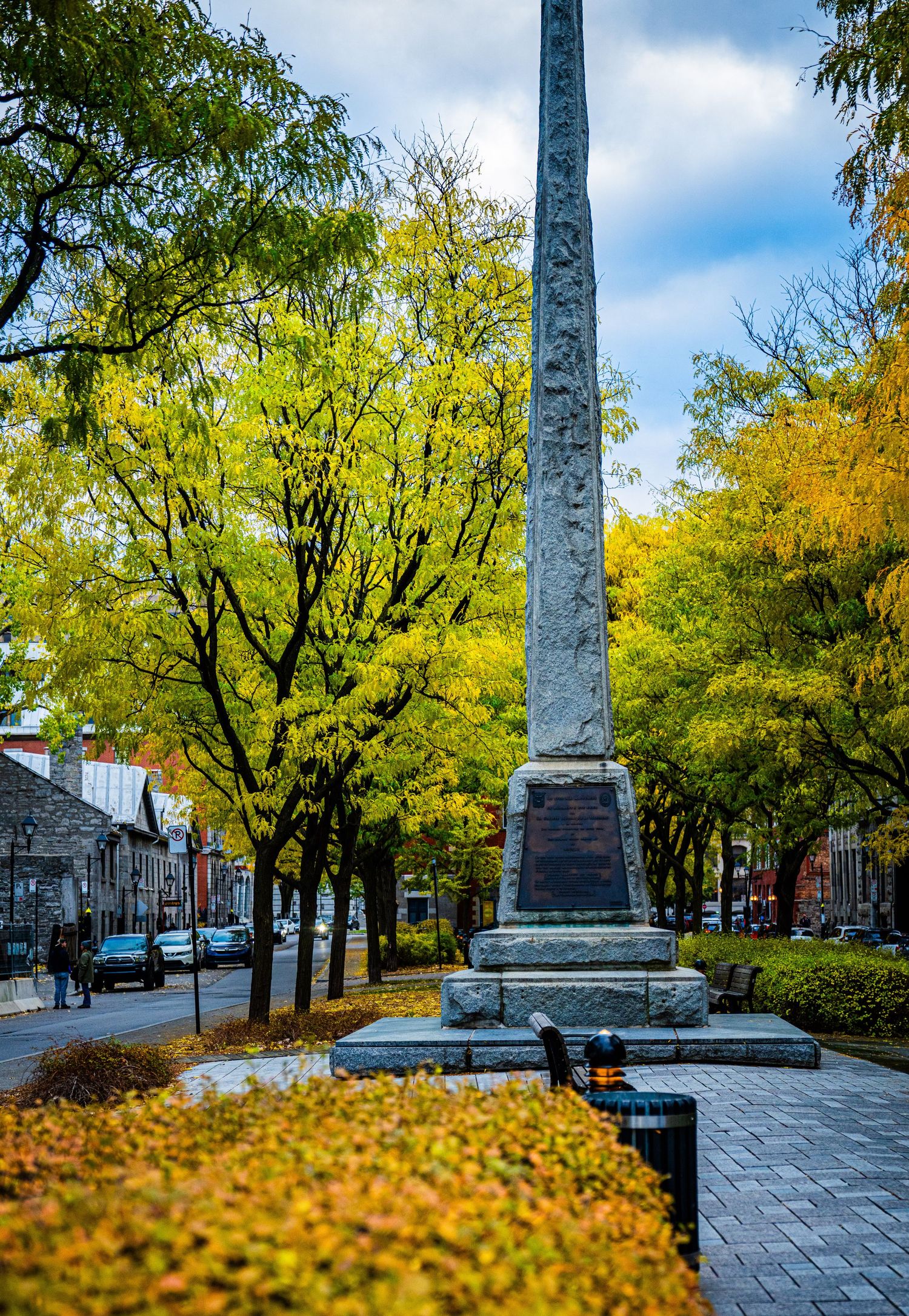 Stone monument stands among vibrant yellow autumn trees in a historic town square with brick buildings in background.