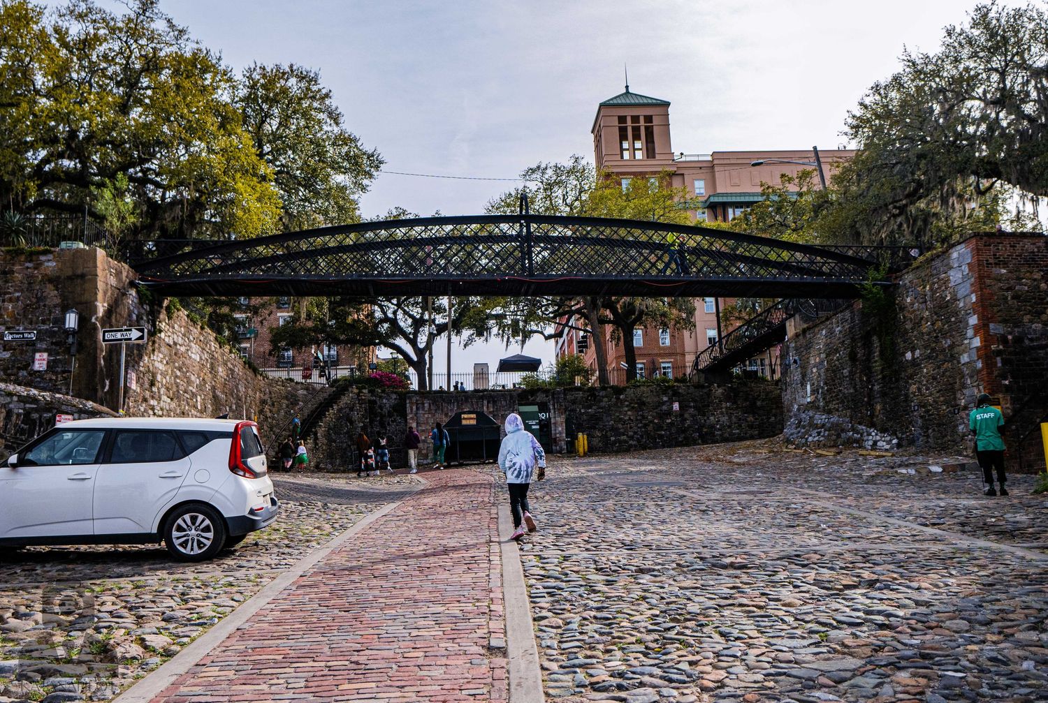 Cobblestone pathway leads to an iron footbridge with historic buildings and autumn trees in the background.