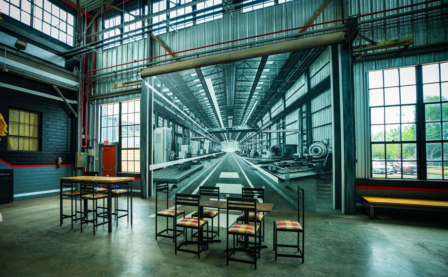 Industrial brewery taproom with vintage factory mural, wooden bar tables, and chairs in a converted warehouse space.
