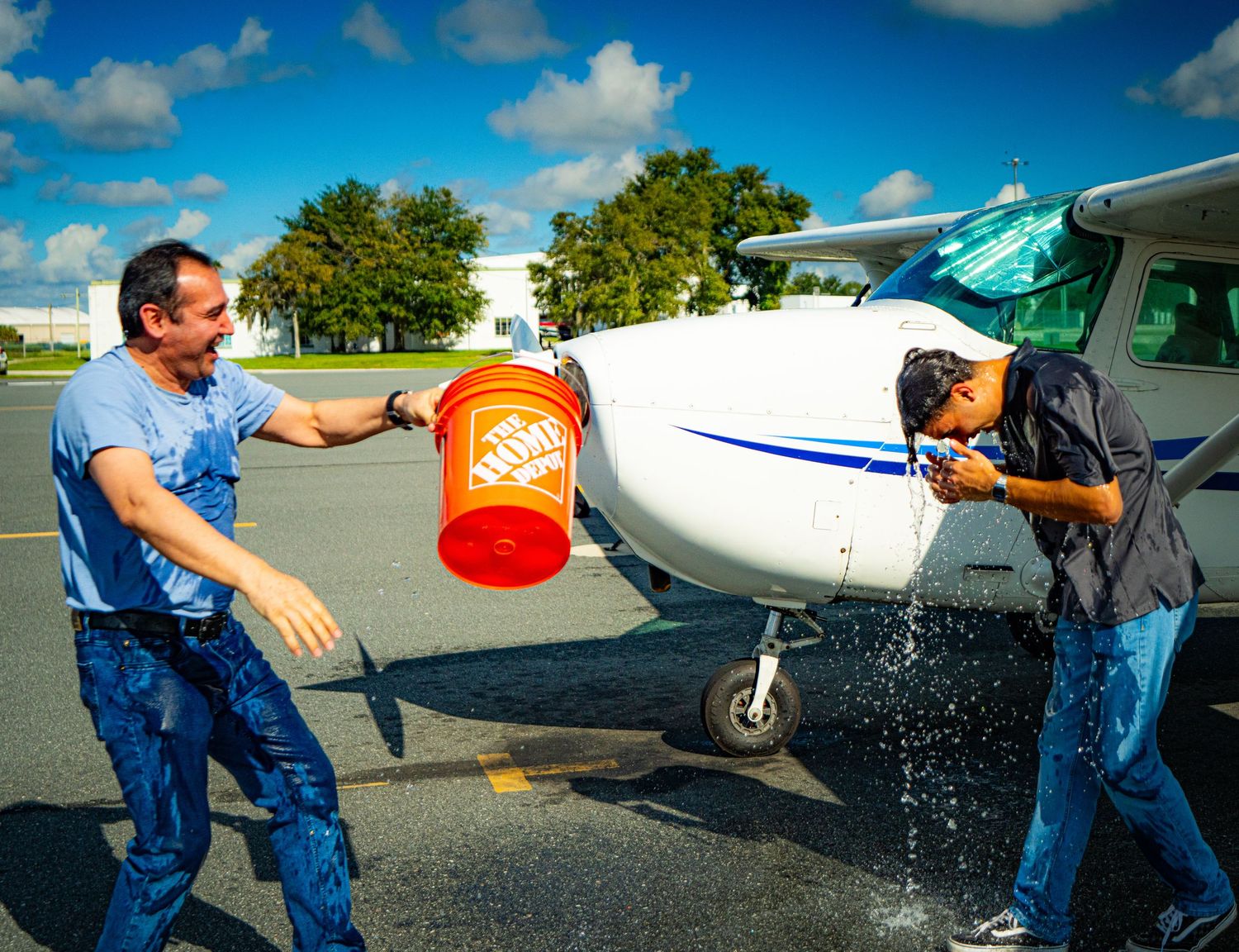 Man splashes water from Home Depot bucket on person near small white Cessna airplane on sunny tarmac.