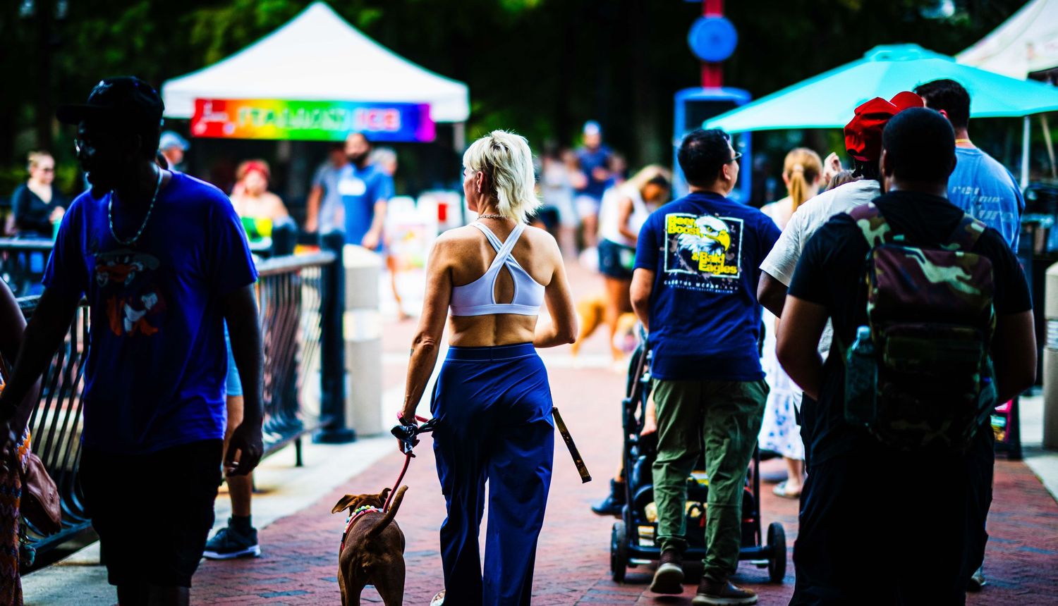 People and a dog walking along a brick path at an outdoor festival with colorful tents.