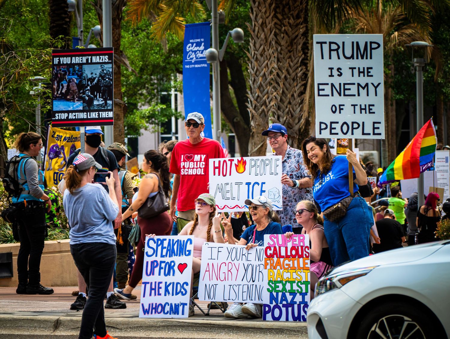 Crowd of demonstrators holding protest signs at a political rally with palm trees in the background.