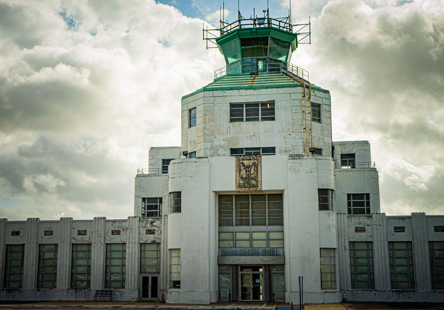Art Deco airport control tower with green roof under dramatic cloudy sky, featuring ornate facade and large windows.
