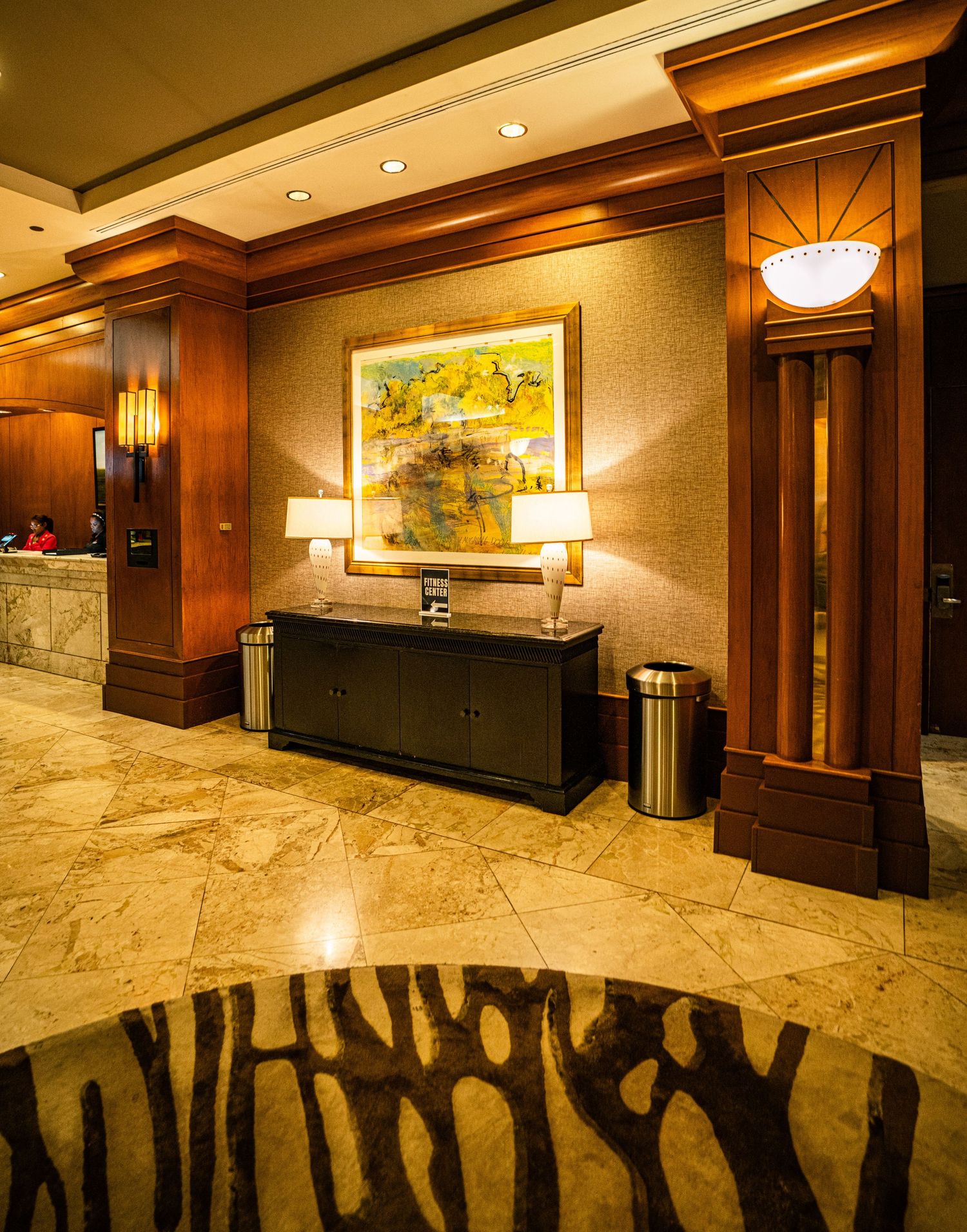 Elegant hotel lobby with marble floors, wood columns, console table with lamps, and colorful abstract wall art.