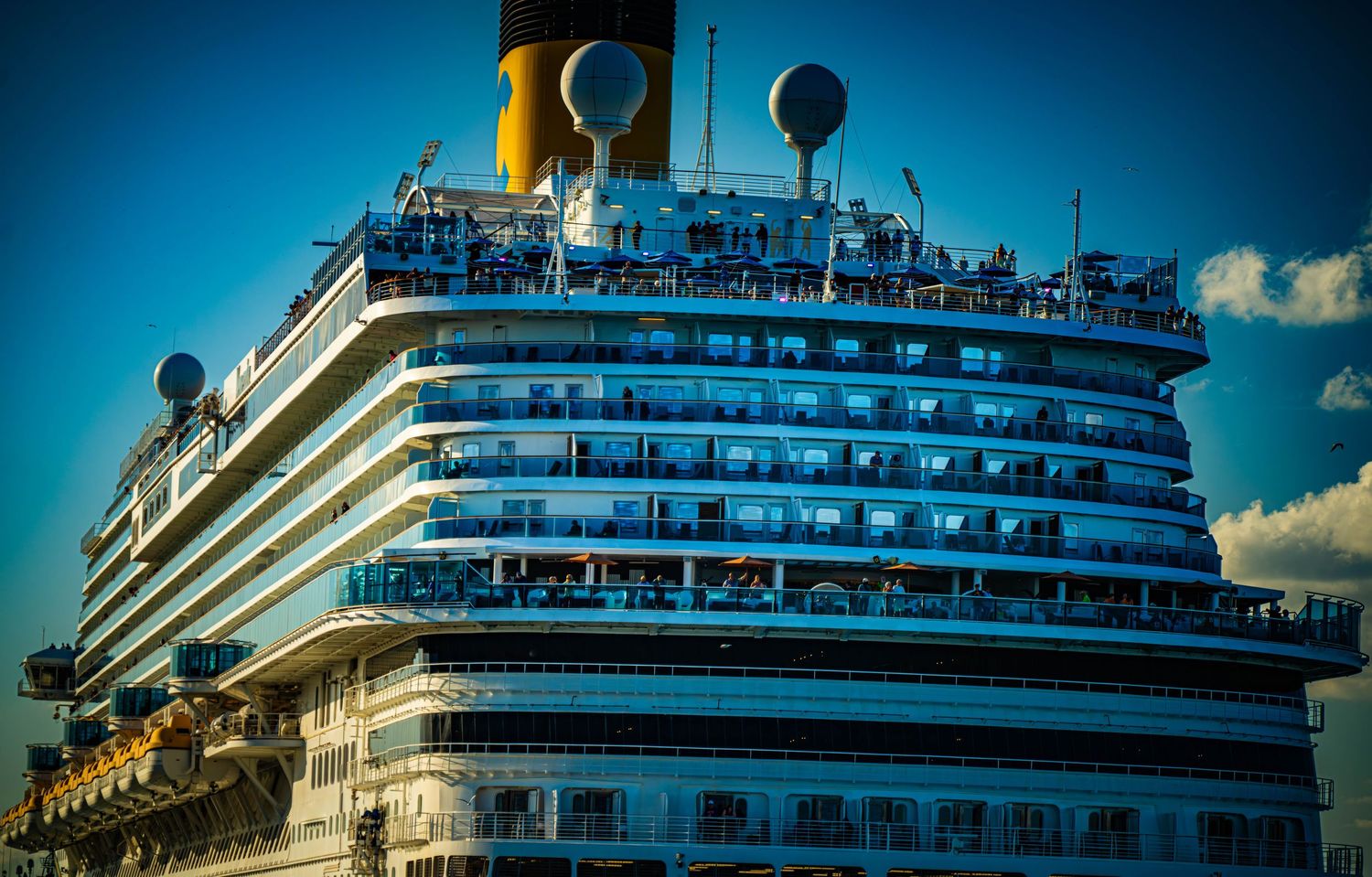 Large cruise ship with yellow funnels and multiple decks photographed against a blue sky with dramatic lighting.