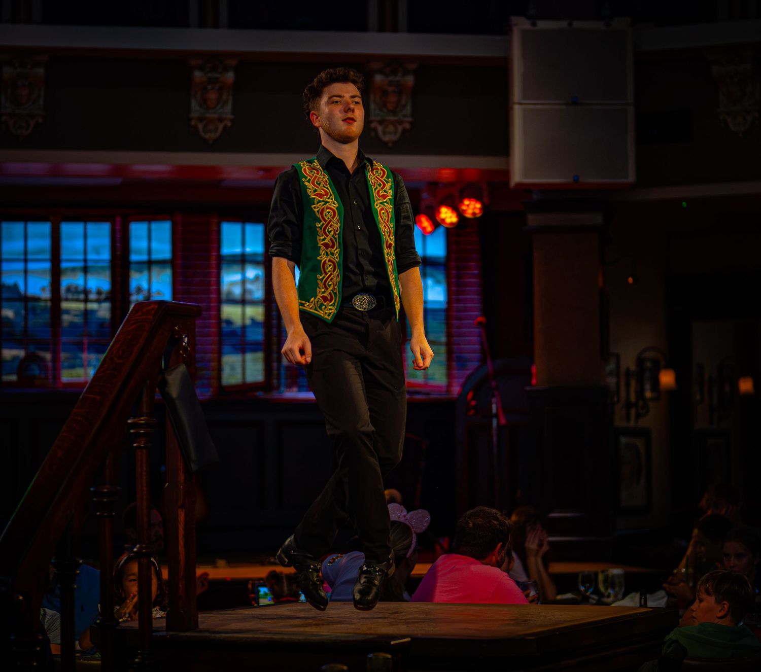 Young male Irish dancer performing on stage in a decorative vest under colorful venue lights.