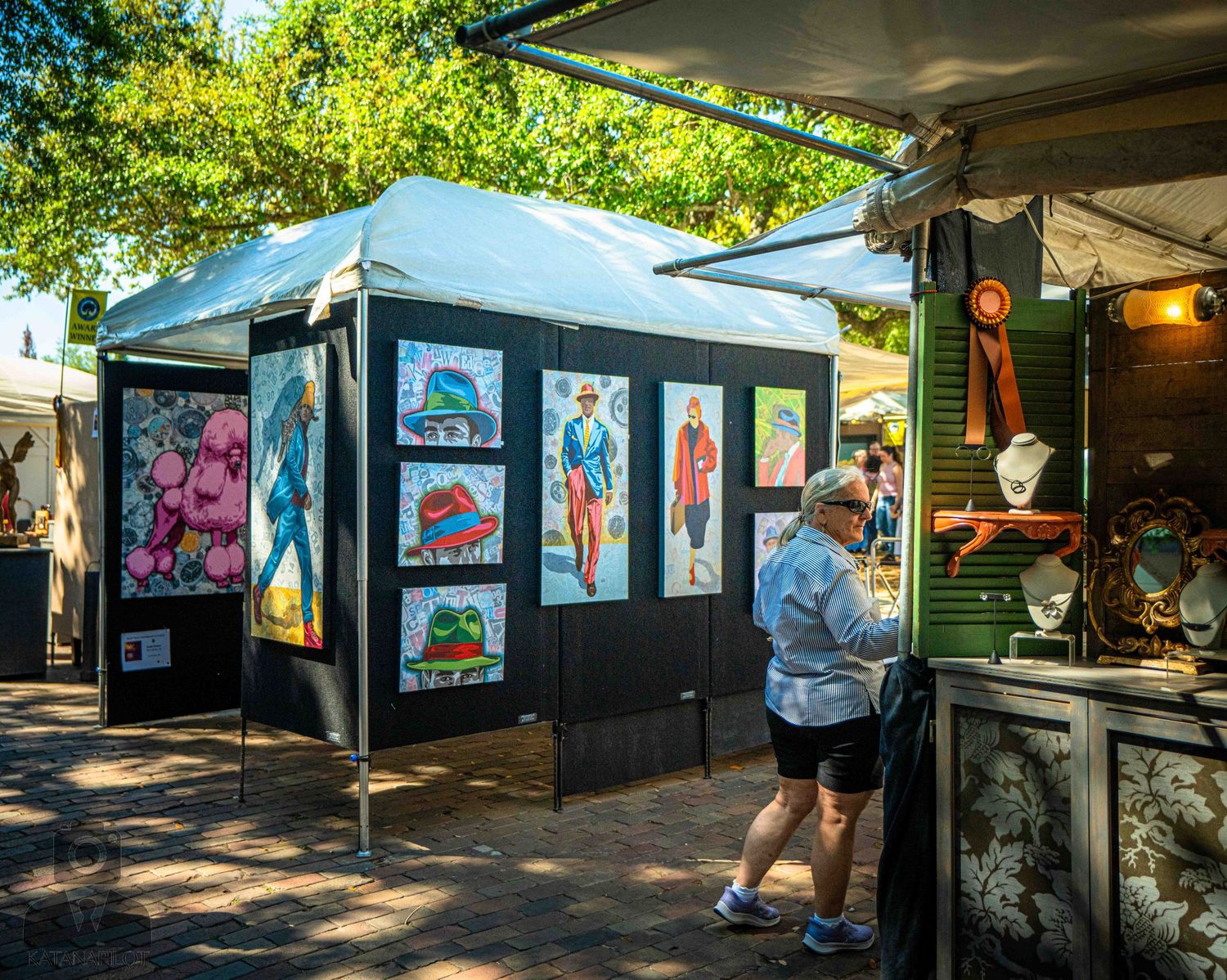 Art exhibition booths displaying colorful artwork under white canopies with visitors walking through.