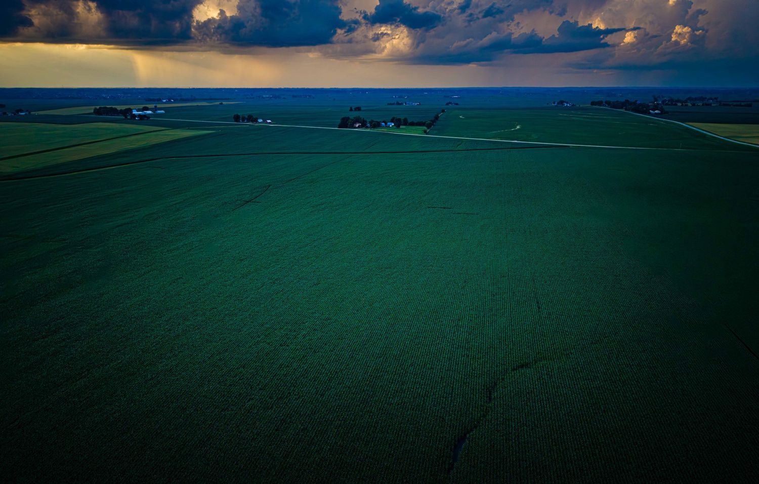 Dramatic aerial view of vast green farmland under stormy skies with dark clouds and rays of light at horizon.