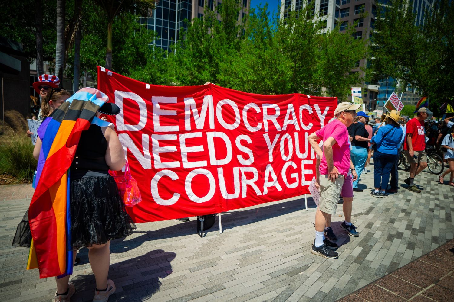 Large red banners with text about democracy and courage are displayed at an outdoor protest rally.