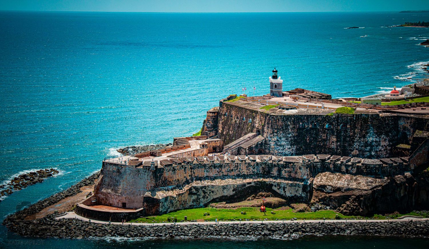 Aerial panoramic view of a coastal fortress with stone walls and grassy areas overlooking turquoise waters.