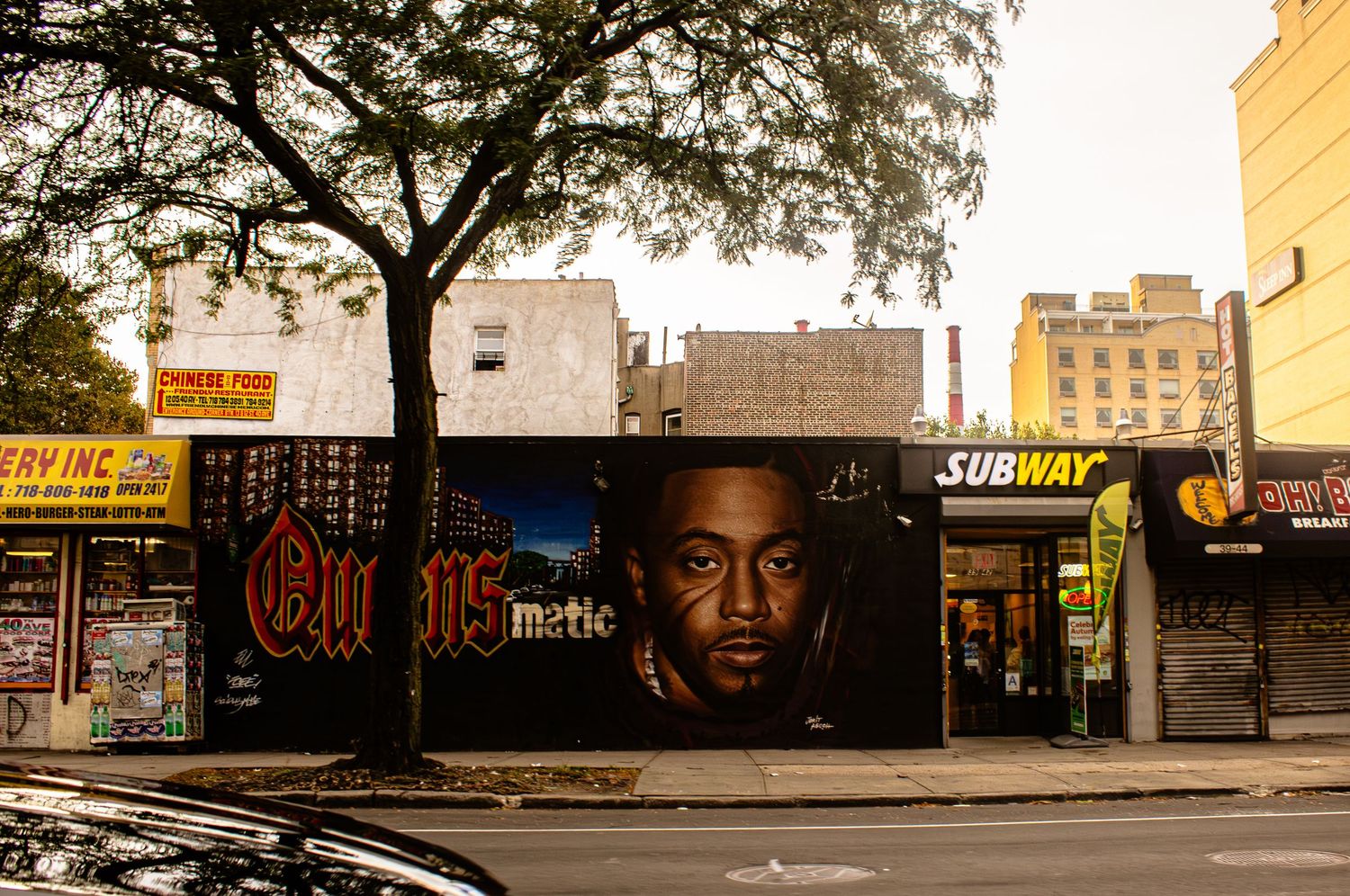 Large street art mural adorns wall of urban storefront next to Subway restaurant and tree in city setting.