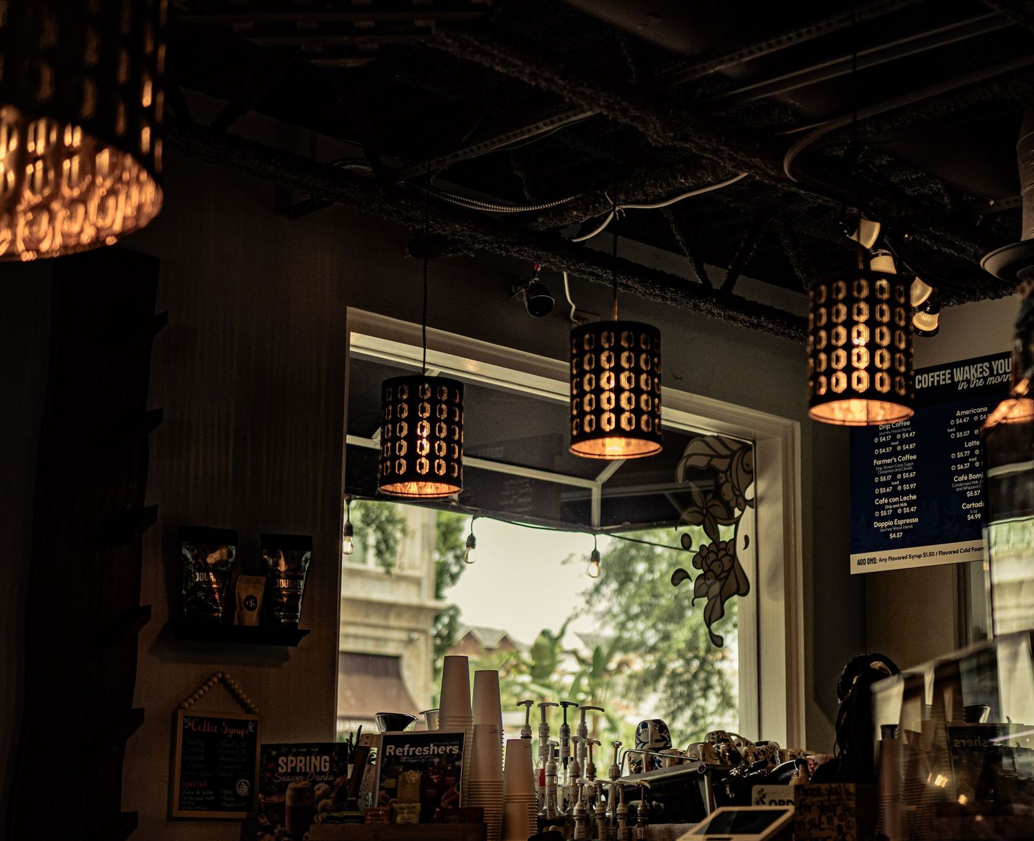 Cozy cafe interior with ornate hanging pendant lights, a menu board, and a bright window view of trees outside.