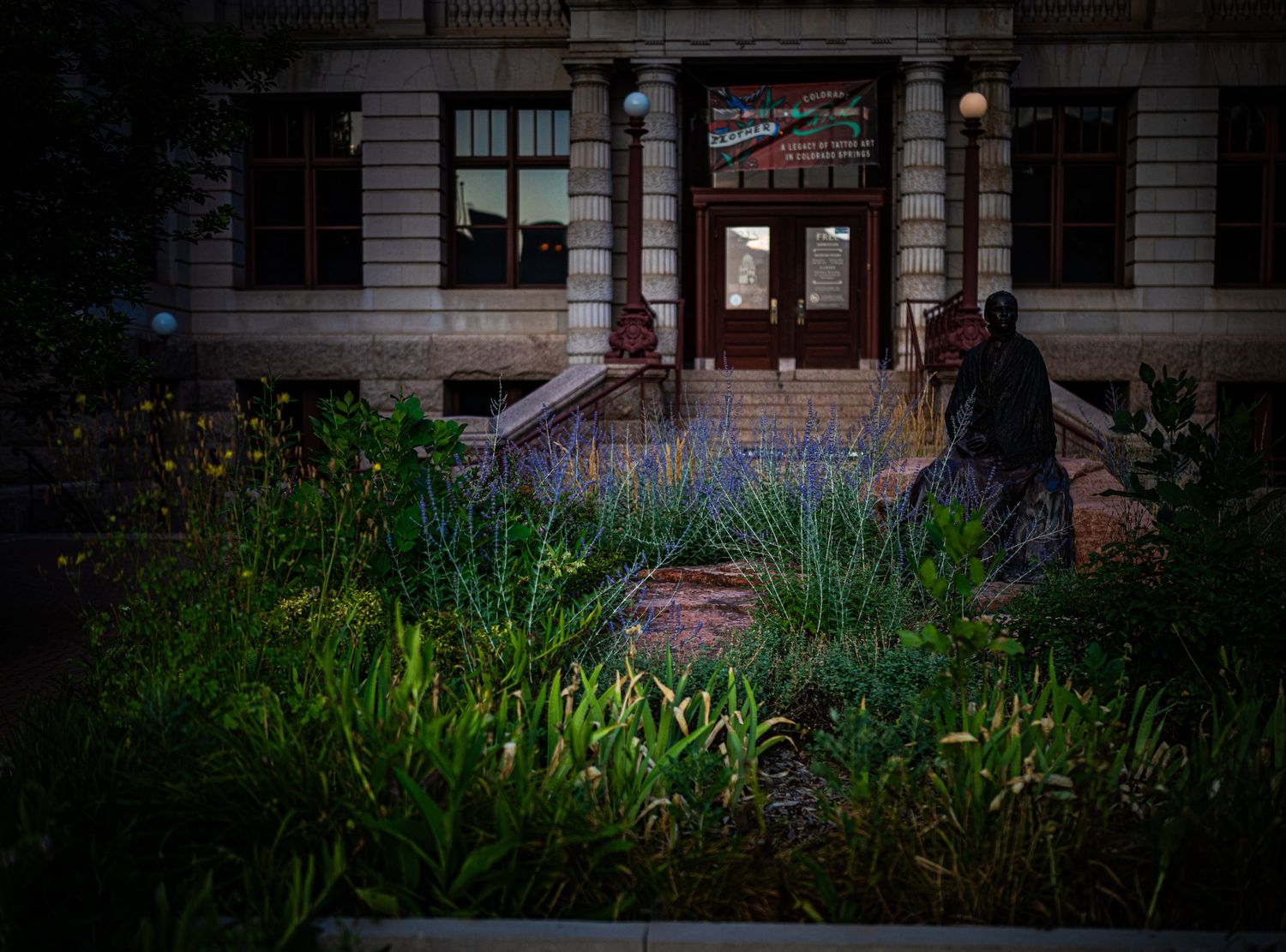 Lush garden with blue flowers and greenery in front of a historic stone building entrance with wooden doors.
