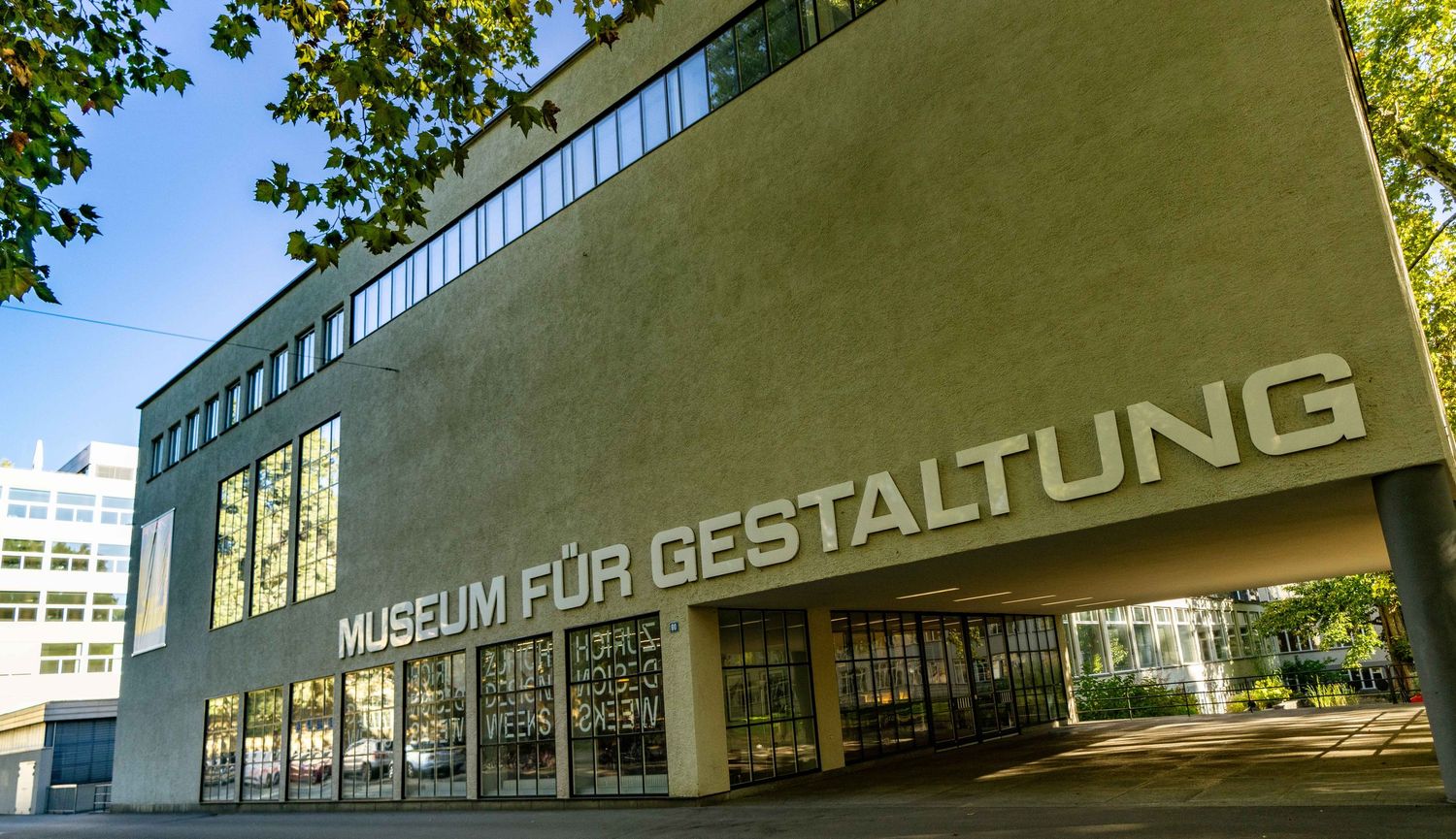 Modernist concrete facade of the Museum für Gestaltung with large windows and bold lettering, framed by green leaves under a blue sky.