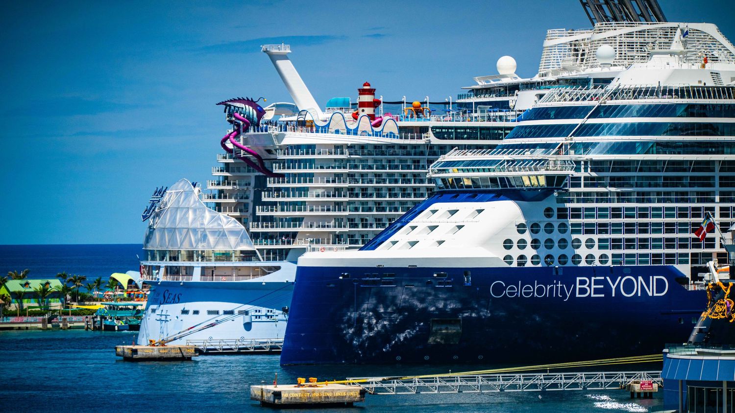Celebrity Beyond cruise ship docked at a tropical port alongside another large cruise ship under a blue sky.