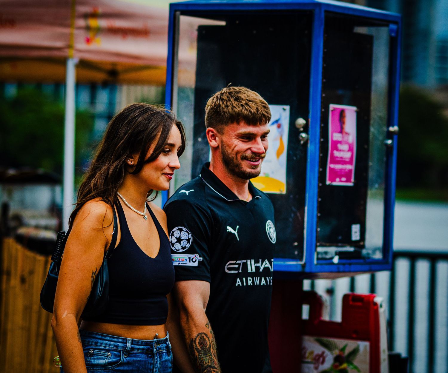 A young couple smiling outdoors, man wearing a Manchester City Etihad Airways jersey, woman in a black crop top.