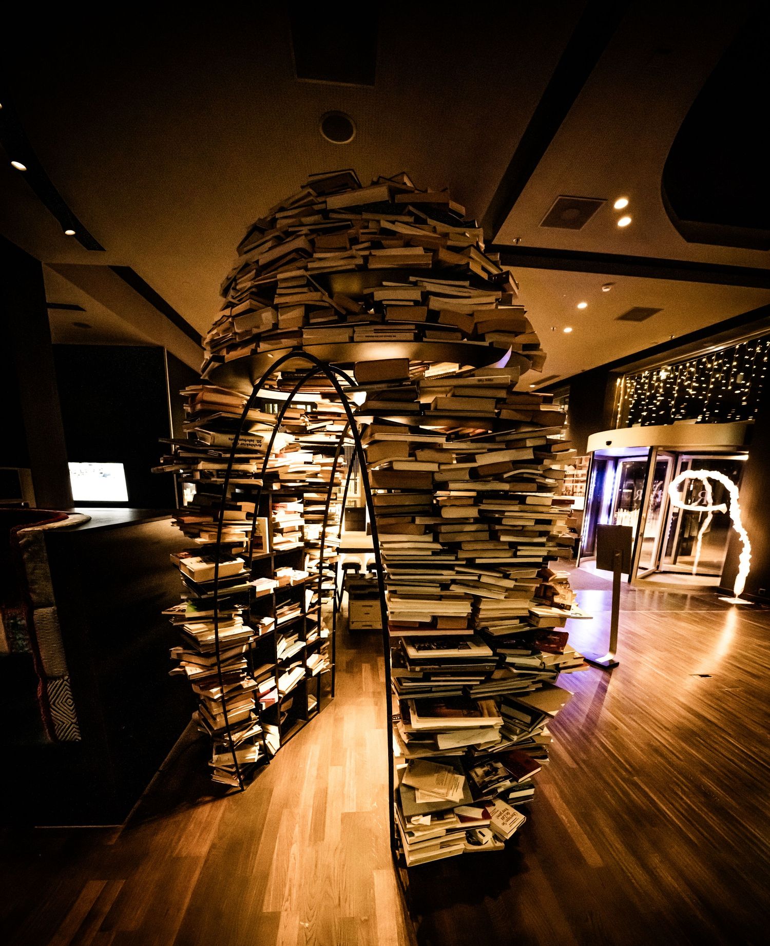 Artistic installation of stacked books creating an archway tunnel in a dimly lit gallery space.
