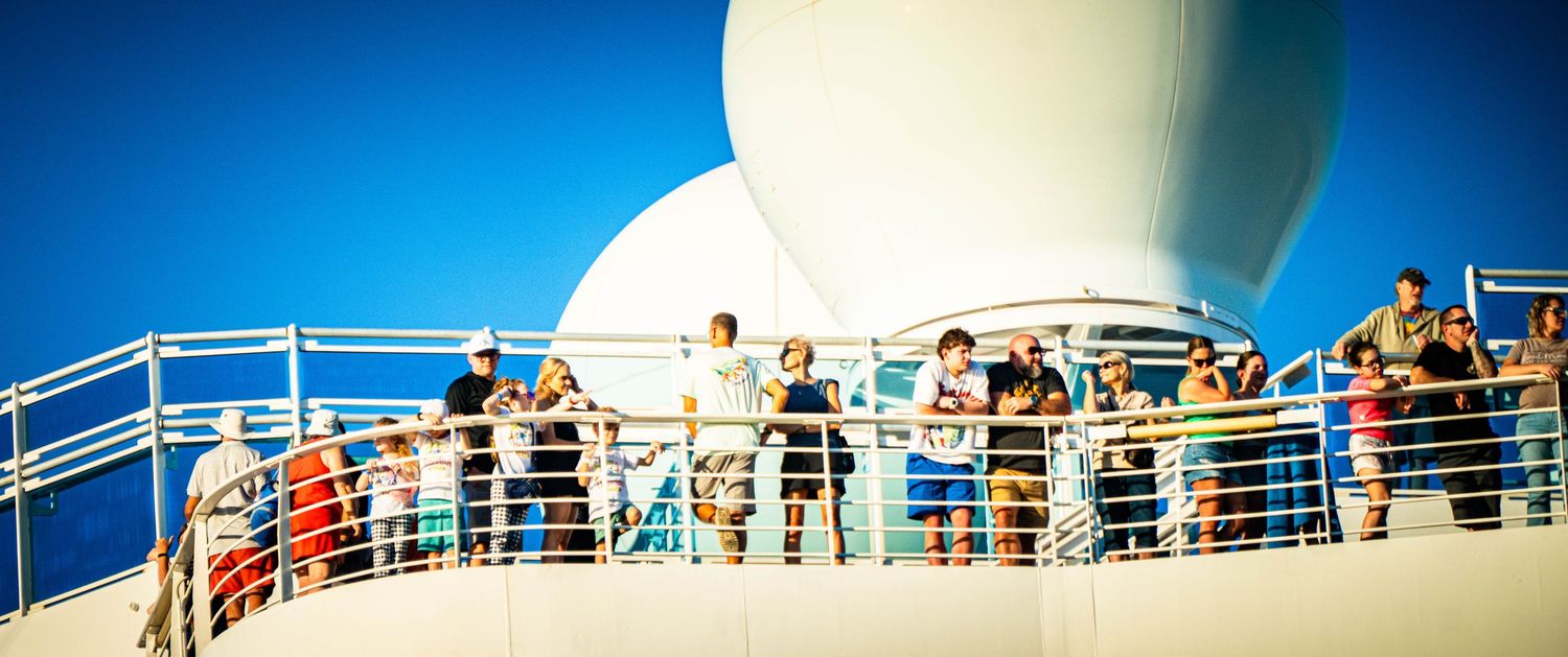 Passengers line the railing of a cruise ship deck against a bright blue sky on a sunny day.