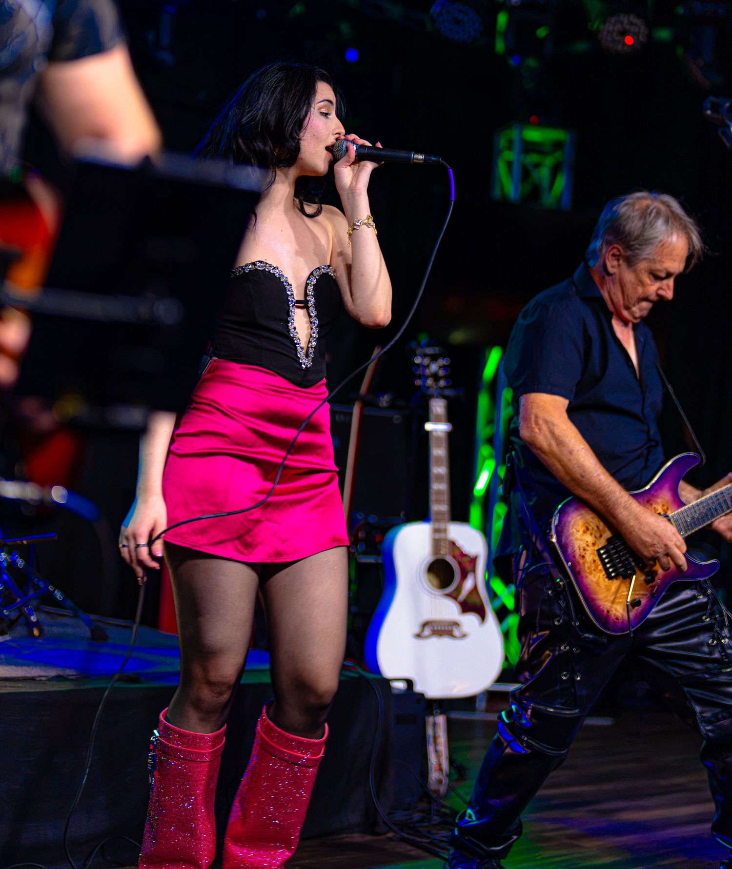 Female singer in pink mini skirt and red boots performs on stage with guitarist playing electric guitar behind her.