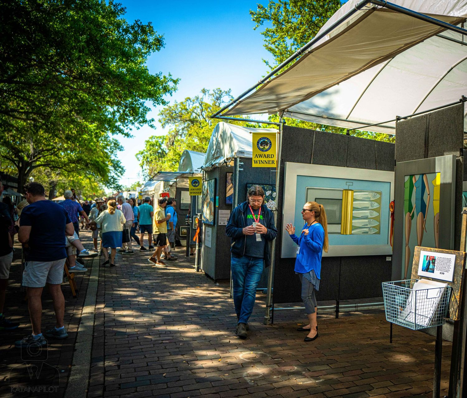 People browse outdoor vendor stalls and displays at a sunny marketplace with trees providing shade overhead.