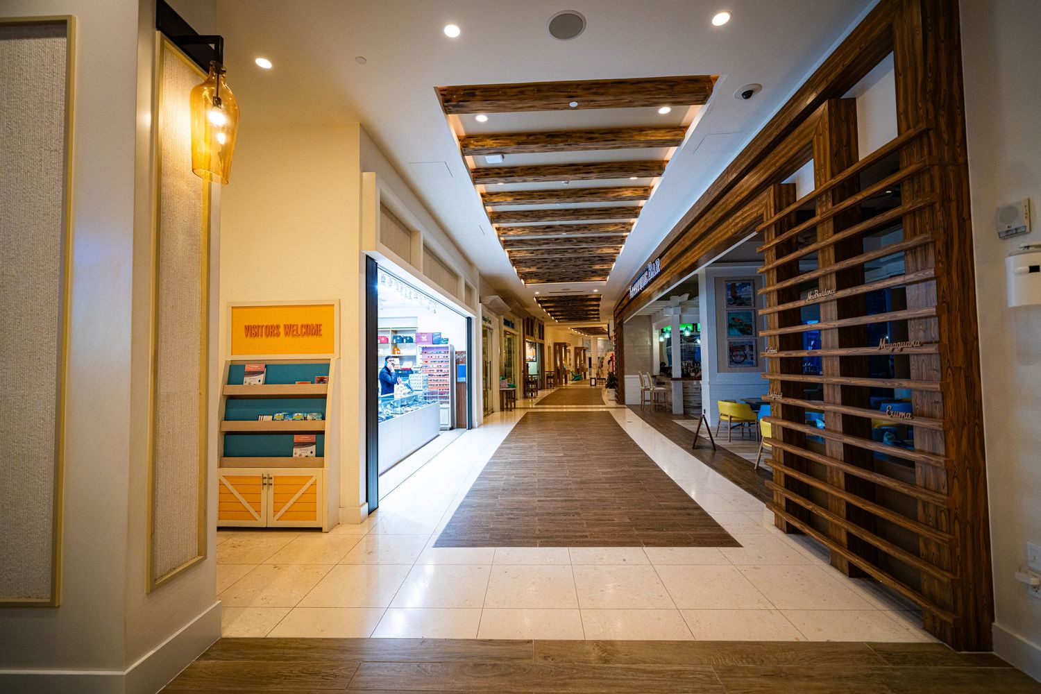 Modern shopping mall corridor with wooden ceiling beams, hardwood floors, and retail storefronts in a bright interior.