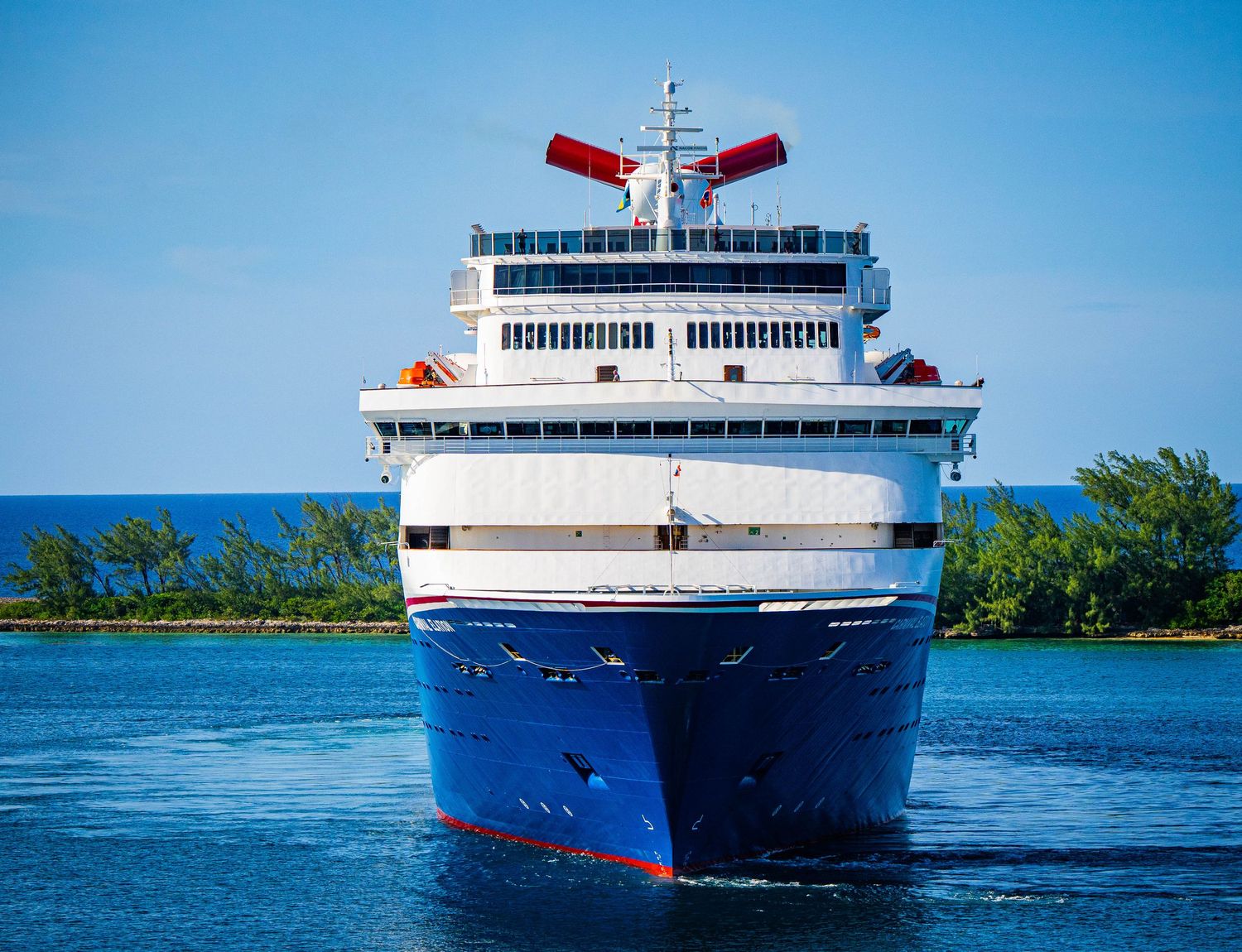 A large Carnival cruise ship with a blue hull approaches port on calm blue waters surrounded by lush green trees.