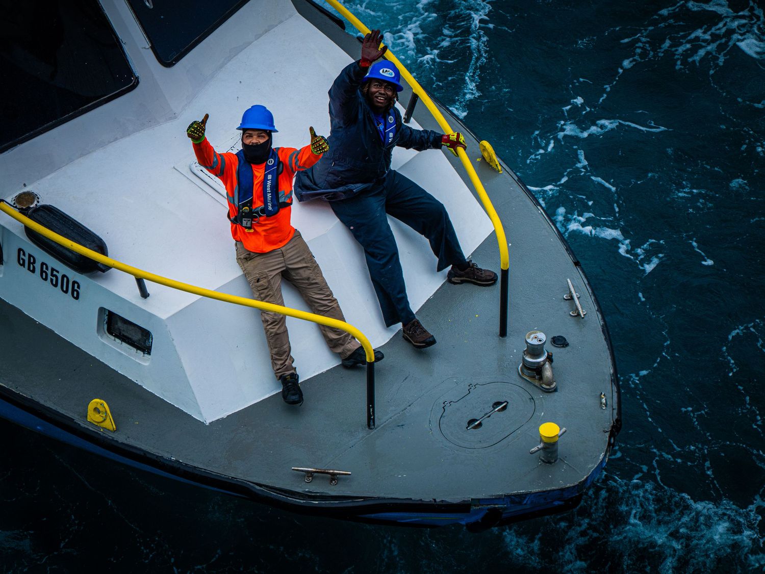 Two workers in safety gear stand on the bow of a vessel surrounded by blue ocean waters.