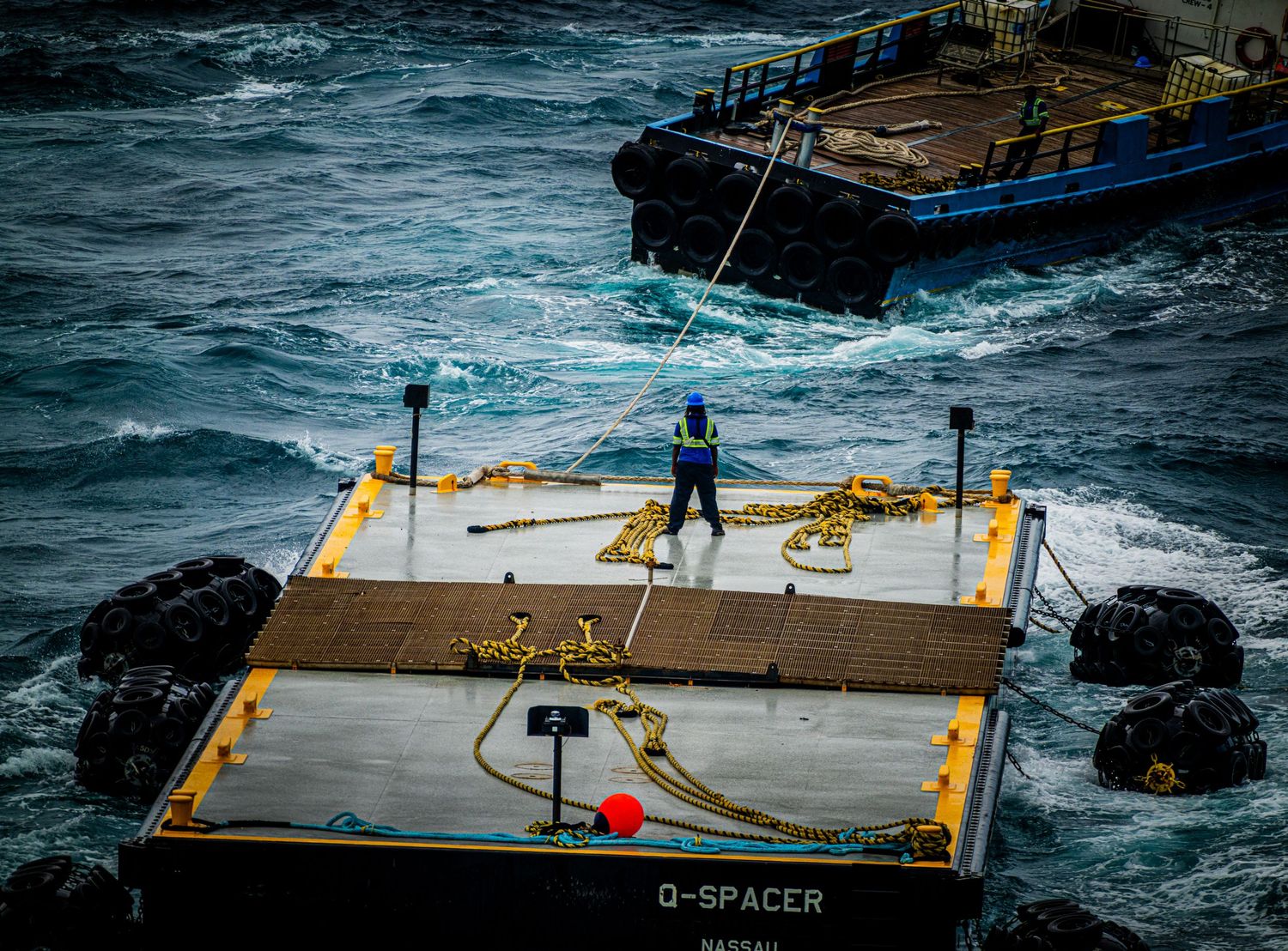 Worker on a D-Spacer floating platform in rough seas with a barge in the background.