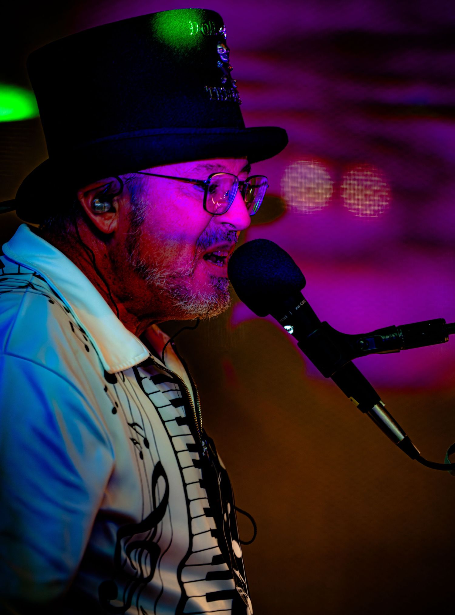 Performer in a hat and piano-themed shirt singing into a microphone under purple and blue stage lights.