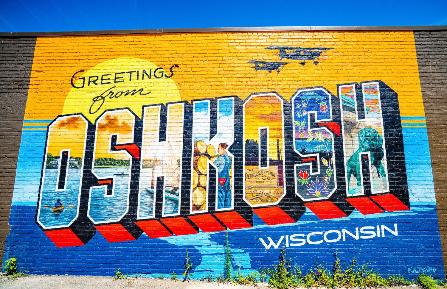 Colorful vintage-style 'Greetings from Oshkosh Wisconsin' mural painted on yellow brick wall with airplane silhouette.