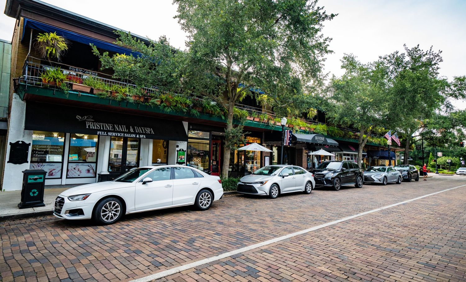 Tree-lined retail street with brick pavers and parked cars along storefronts.