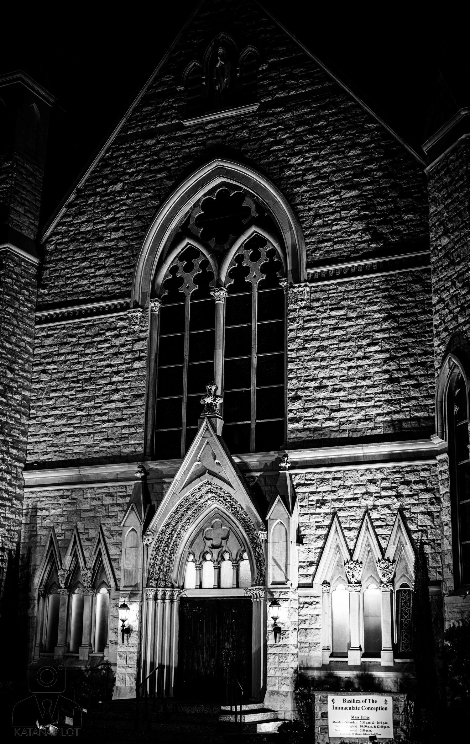 Black and white night photo of a Gothic stone church facade with arched windows and illuminated entrance doors.