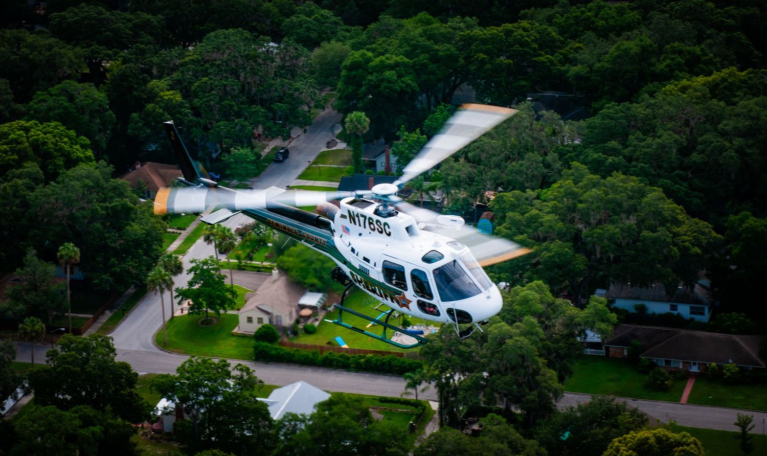 White helicopter hovering over parking lot surrounded by lush green trees.