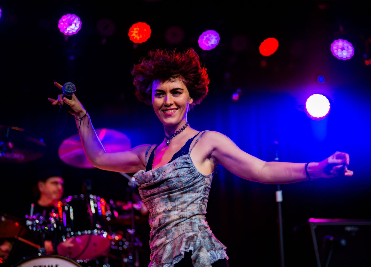 Female performer with red curly hair singing on stage with colorful concert lights behind her.