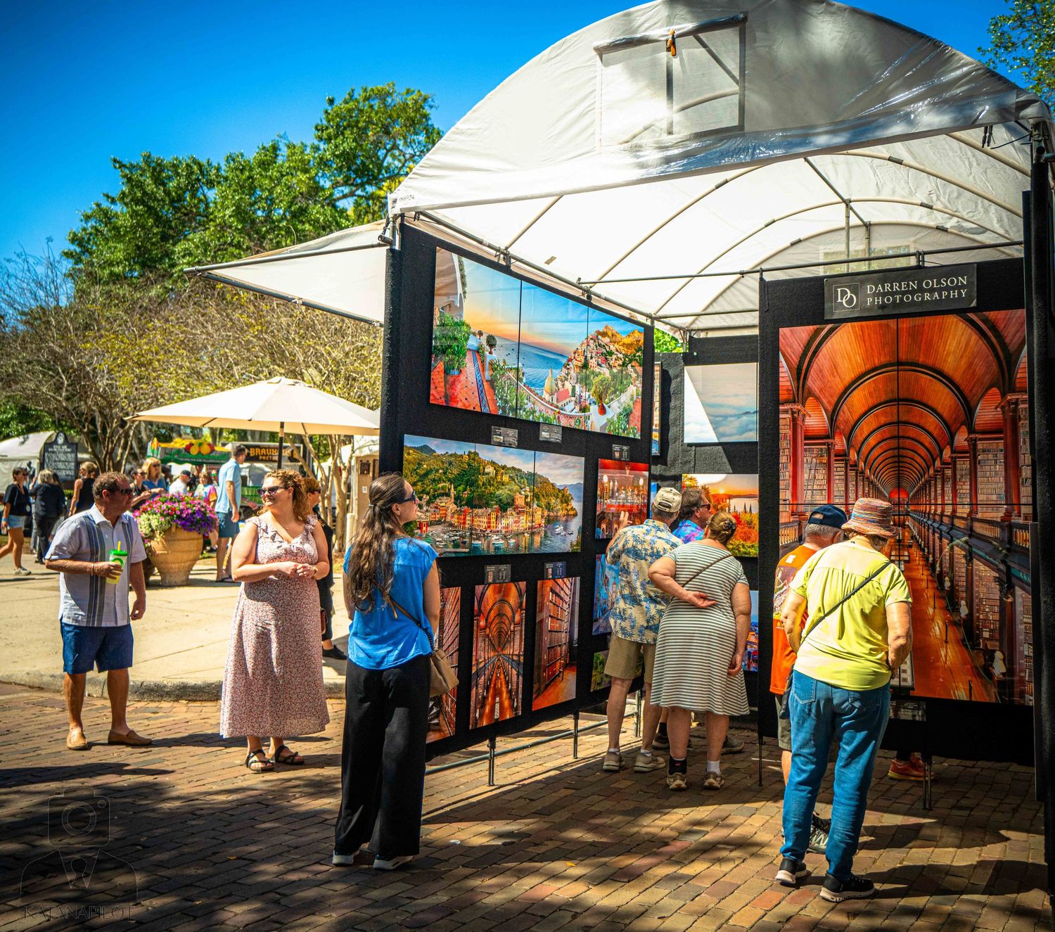 Covered walkway with white canopy shelters people browsing vendor stalls at an outdoor market during daytime.