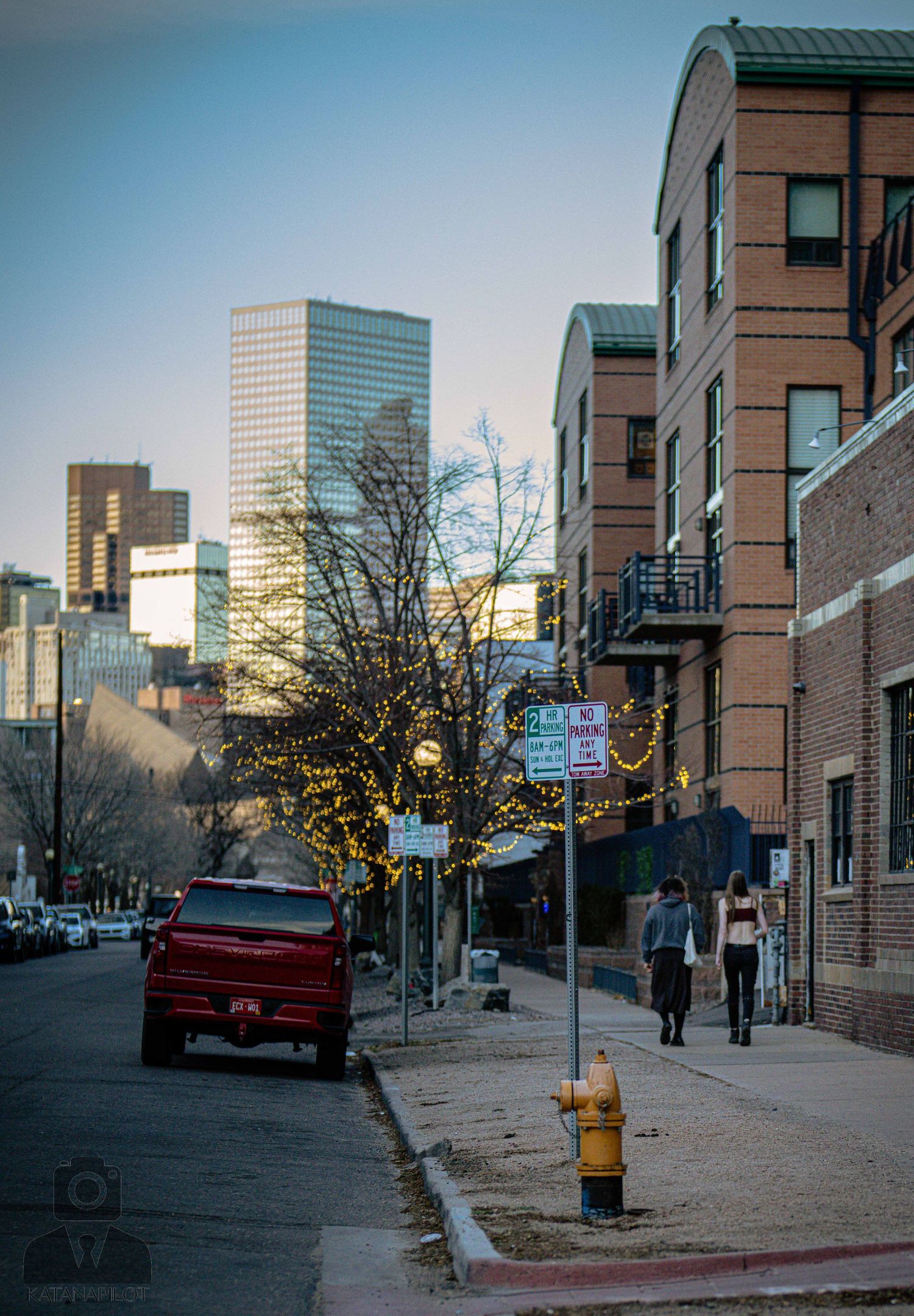 Urban street scene in downtown Denver showing brick buildings, parked cars, and high-rise office towers in the background.