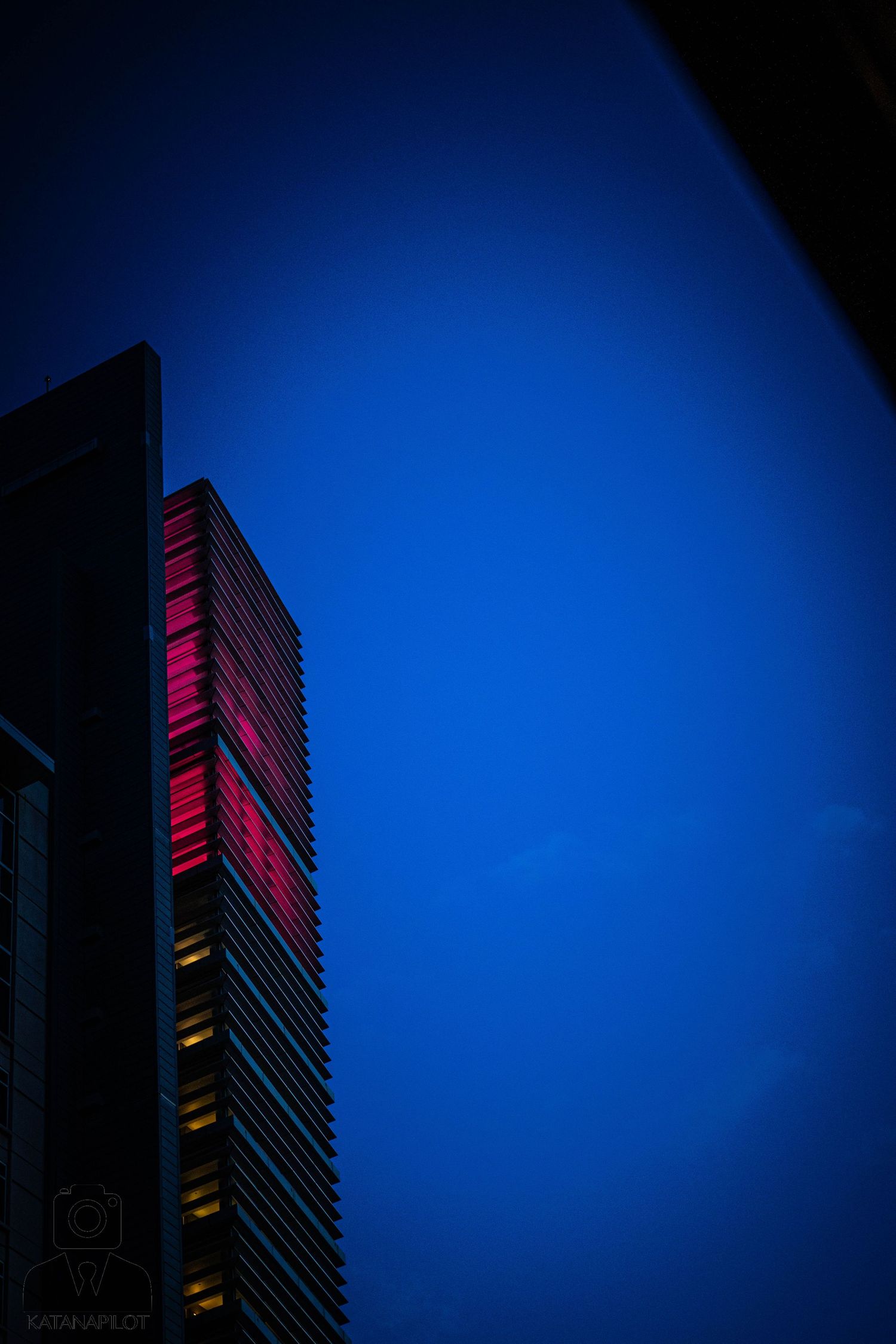 Modern high-rise building illuminated with pink and red lights against a deep blue night sky.