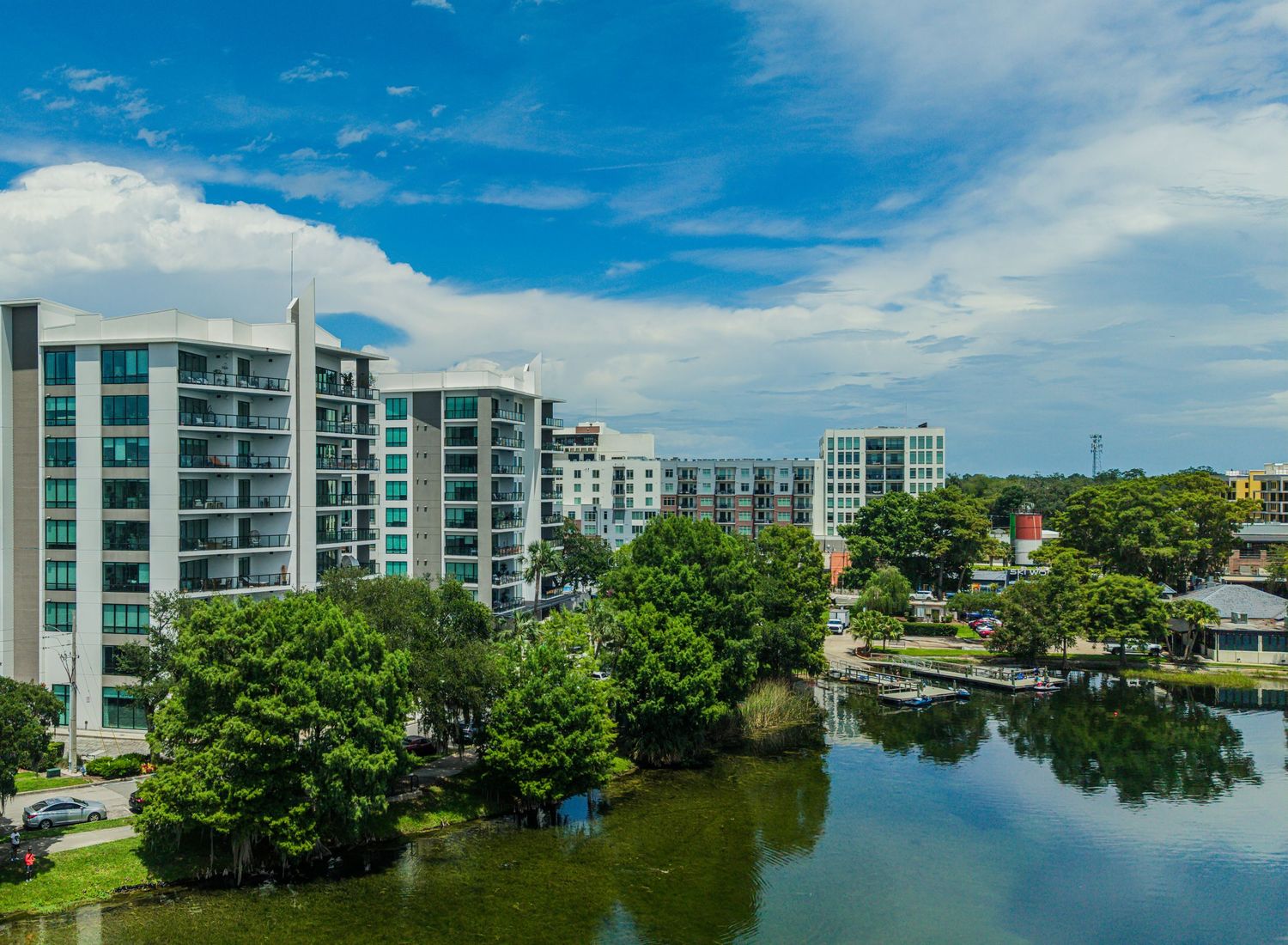 Waterfront condominiums and lush green trees reflect along a calm urban canal under a bright blue sky.